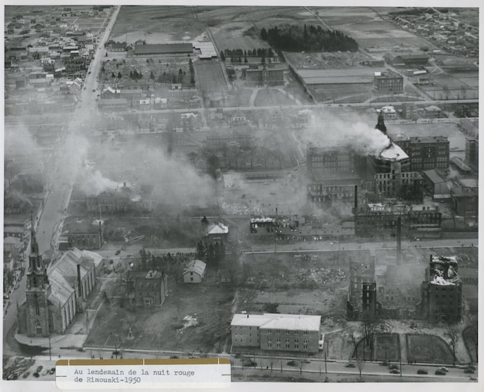 Vue aérienne du centre-ville de Rimouski au lendemain de l'incendie.  De la fumée s'échappe de nombreux édifices. 